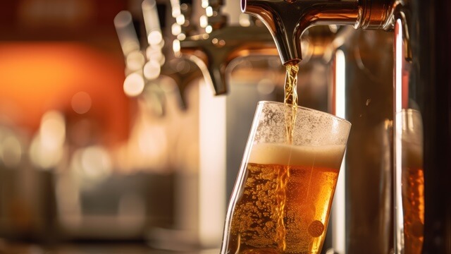 Beer pouring from a tap into a tilted glass, forming a frothy head with a warm, blurred bar background